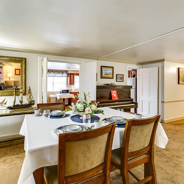 Dining room with a set table, floral centerpiece, mirror, and piano, leading to a kitchen.