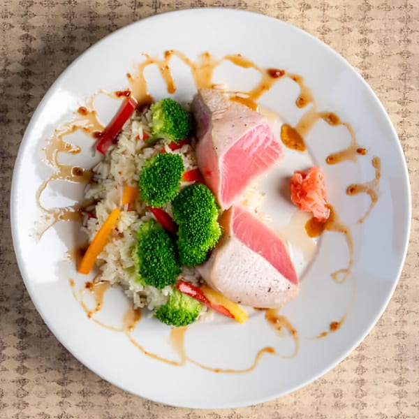 Seared tuna with rice, broccoli, bell peppers, and ginger, drizzled with sauce on a white plate.