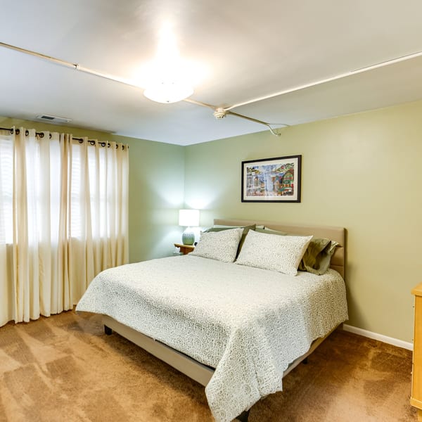A cozy bedroom with a queen-sized bed, light green walls, and a large window with sheer curtains.
